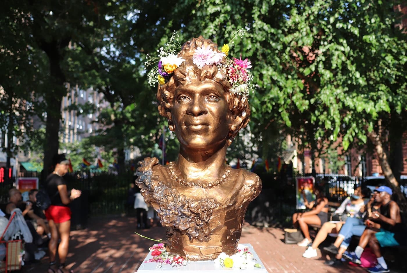 A bronze bust of Marsha P. Johnson in a park, with fresh flowers tucked into her hair and laid at its base, people sitting on benches behind.